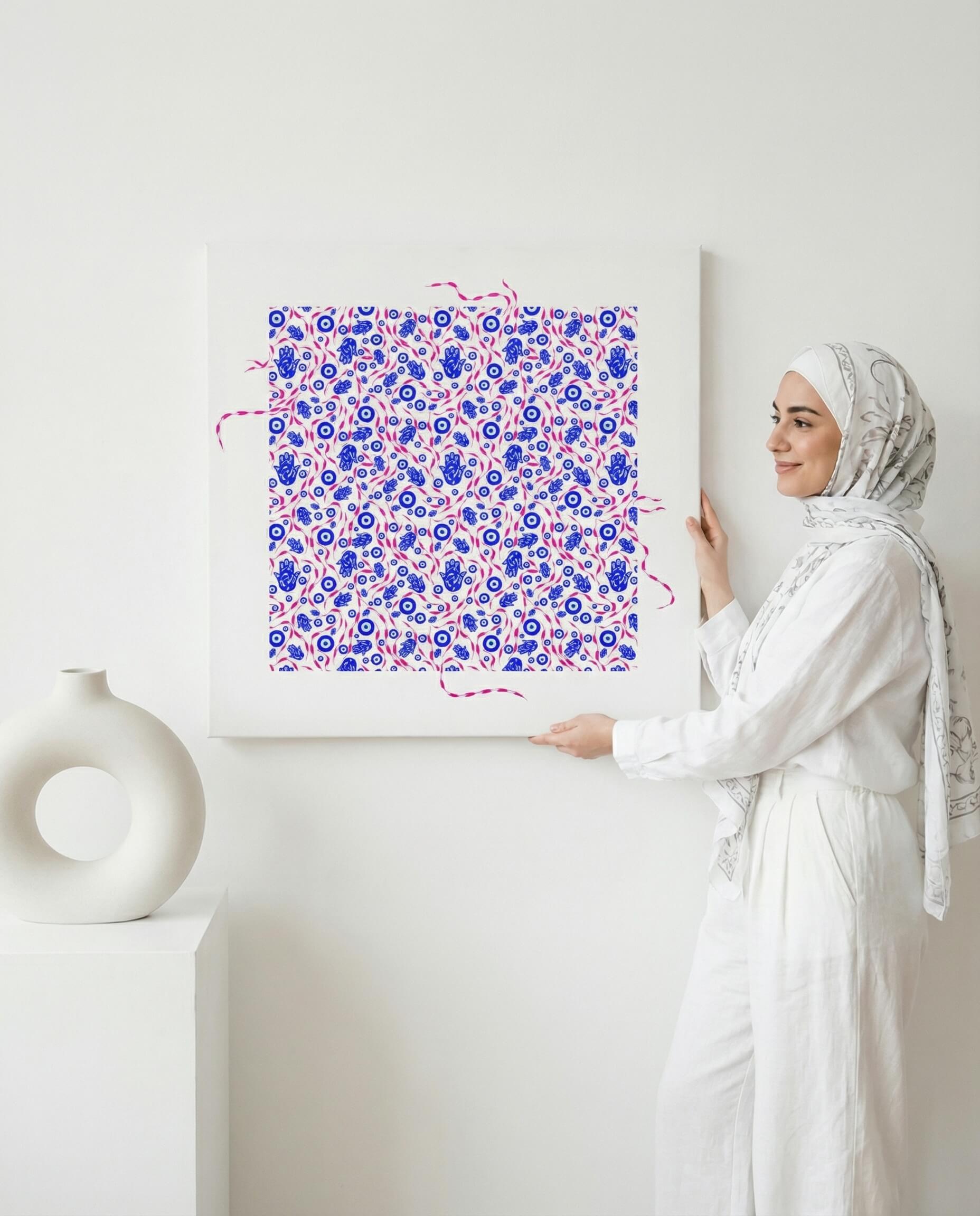 Woman in hijab holding a framed islamic abstract art piece with blue and pink patterns on a white wall.