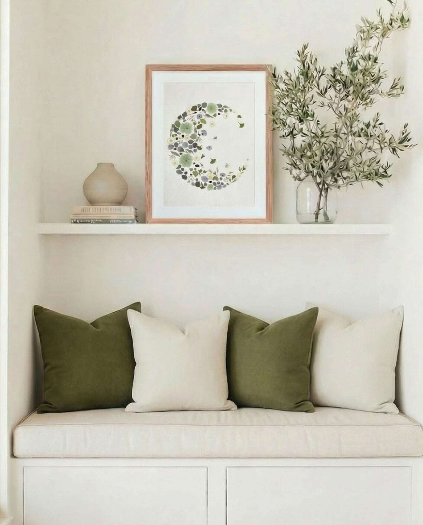 Framed fading moon artwork made from dried flowers displayed on a shelf above a built-in bench in a light, neutral interior.