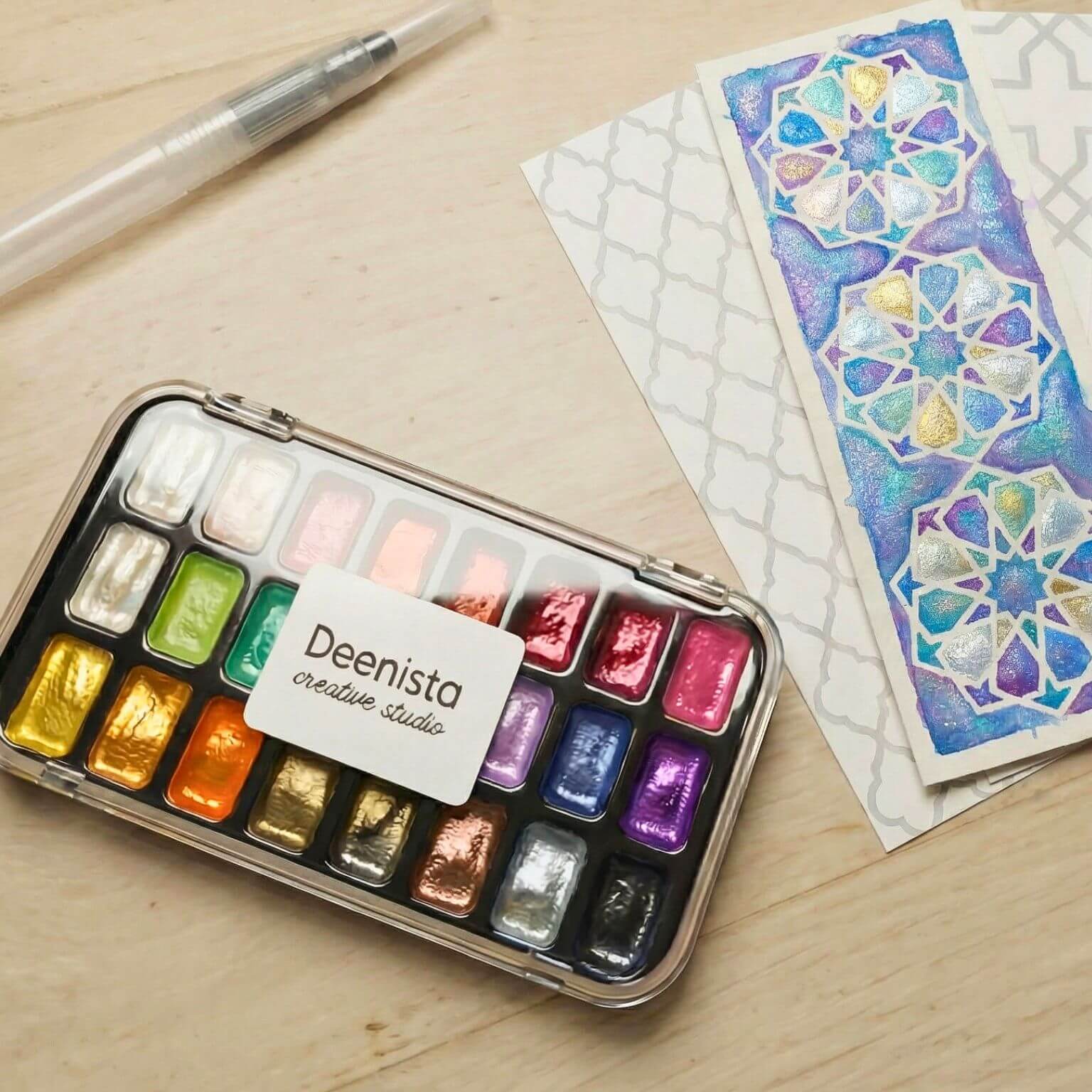 Deenista Islamic shimmer watercolor kit with refillable water brush, painted Islamic geometric bookmark, and blank bookmark cards on a wooden surface